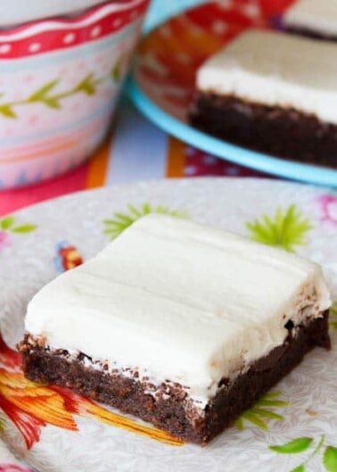 A piece of frosted brownie on a colorful floral napkin with more brownies and a patterned bowl in the background.