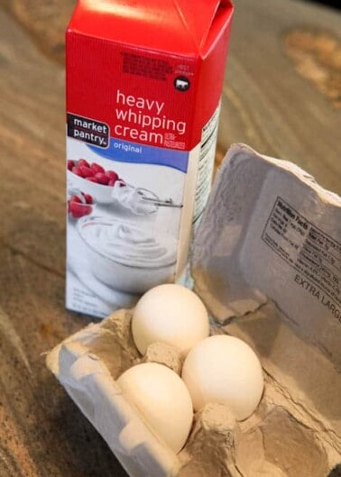 Carton of heavy whipping cream and three eggs on a wooden surface.