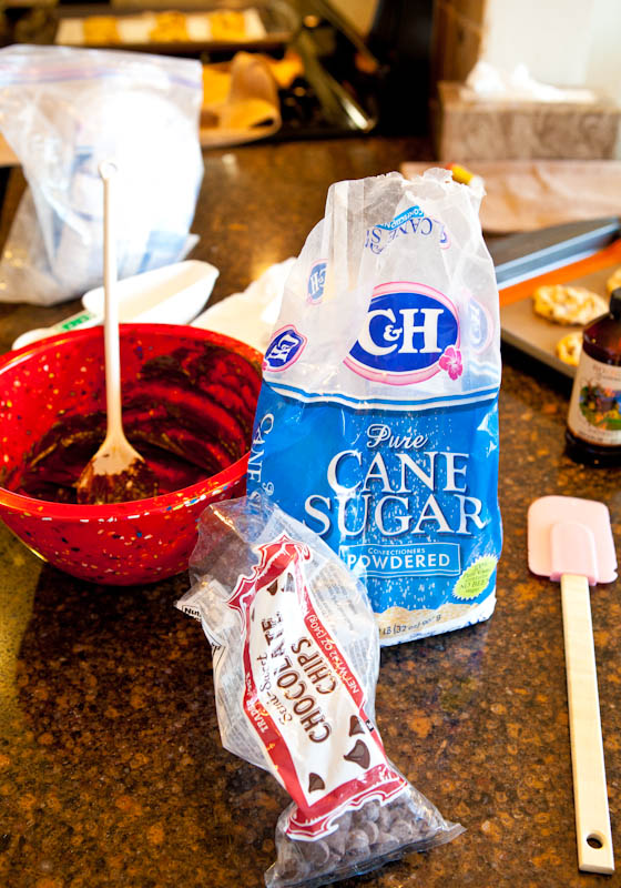 Bag of powdered sugar, red bowl of chocolate and sugar, and bag of chocolate chips with pink spatula