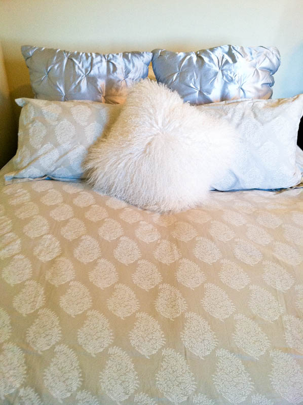 Bed with yellow patterned sheets, silver and white pillows with fuzzy throw pillow