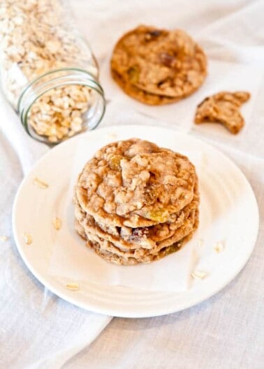 A stack of oatmeal cookies on a white plate with a jar of oats spilling out nearby.