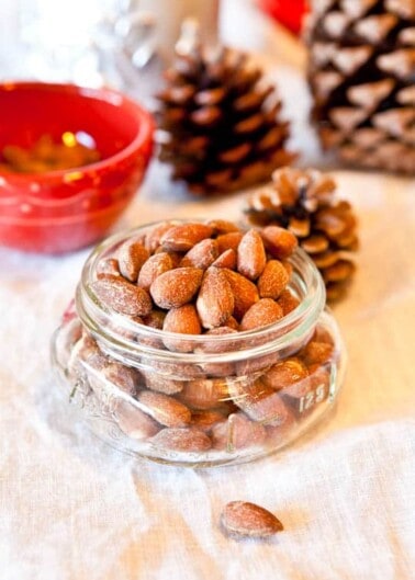 Jar of almonds on a table with pinecones and a bowl in the background.