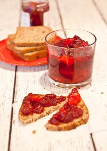 A slice of bread with jam on a napkin, a glass of jam, and additional slices of bread with a jar of jam in the background on a wooden surface.