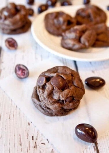 Chocolate cookies with chocolate chips on a white plate, scattered candies nearby on a wooden surface.