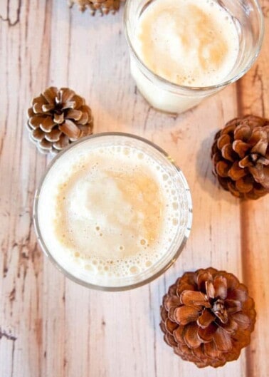 Two glasses of frothy beverage with pine cones on a wooden surface.