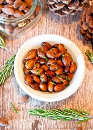 A bowl of seasoned almonds surrounded by rosemary sprigs on a wooden surface.