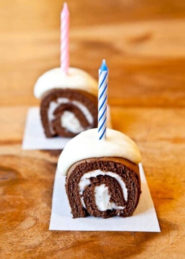 Two chocolate cake rolls with white cream filling and a lit birthday candle in each on a wooden surface.
