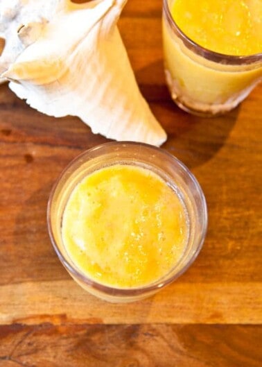 A glass of orange smoothie on a wooden table, with another glass and a seashell in the background.