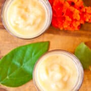 Two glasses of creamy yellow smoothie on a wooden surface with green leaves and orange flowers.