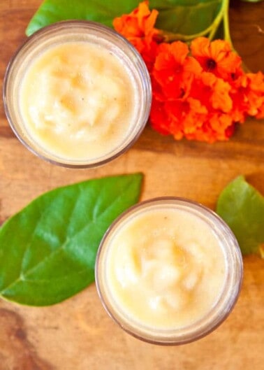 Two glasses of creamy yellow smoothie on a wooden surface with green leaves and orange flowers.