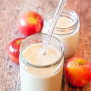 Two jars of apple smoothie with straws on a wooden surface, accompanied by fresh apples.