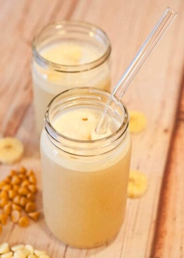 Two jars of banana smoothie with a clear straw, accompanied by banana slices and nuts on a wooden surface.