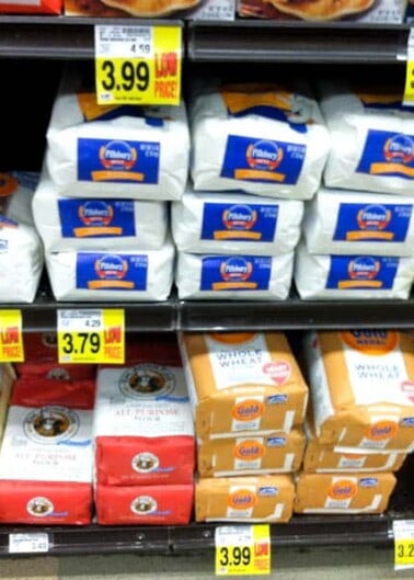 A grocery store shelf stocked with various brands and types of flour, each with a price tag displayed.