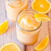 Two jars of orange smoothie with orange slices on a wooden surface.