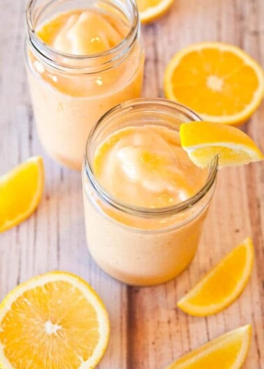 Two jars of orange smoothie with orange slices on a wooden surface.