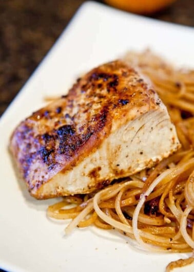 Grilled chicken breast served on top of spaghetti noodles on a white plate.