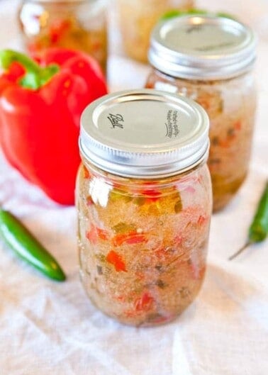 Three jars of homemade salsa with a red bell pepper and a jalapeño pepper in the background.
