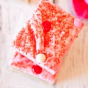 A piece of pink frosted cake with red sprinkles and candy toppings on a wooden surface.
