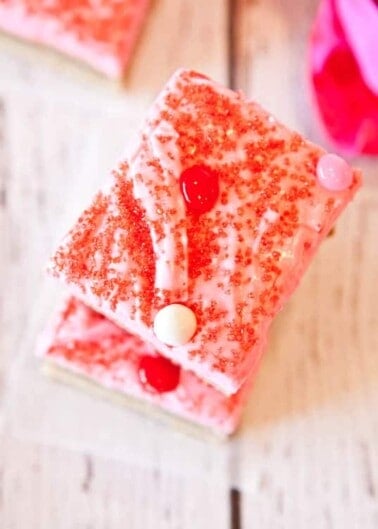 A piece of pink frosted cake with red sprinkles and candy toppings on a wooden surface.