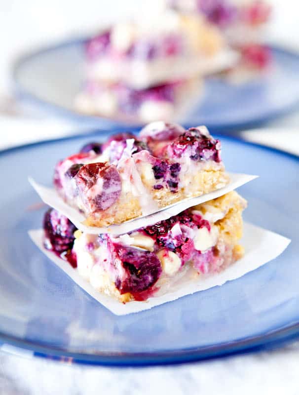 Two berry dessert bars stacked with parchment paper between them on a blue plate, topped with mixed berries and white drizzle.