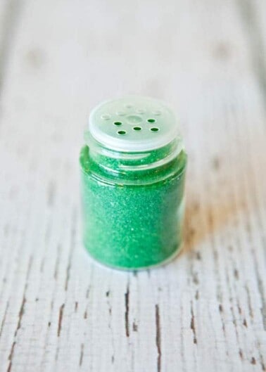 A small jar of green glitter on a wooden surface.