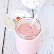 A glass jar filled with pink smoothie and a straw on a white wooden surface with a floral cup and saucer in the background.