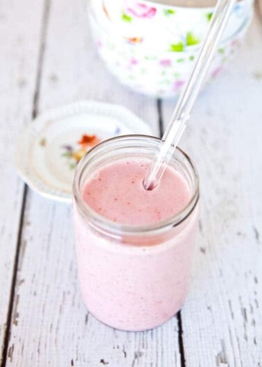 A glass jar filled with pink smoothie and a straw on a white wooden surface with a floral cup and saucer in the background.