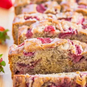 Sliced strawberry banana bread with visible chunks of strawberries on top, placed on a wooden surface. Some whole strawberries are visible in the background.