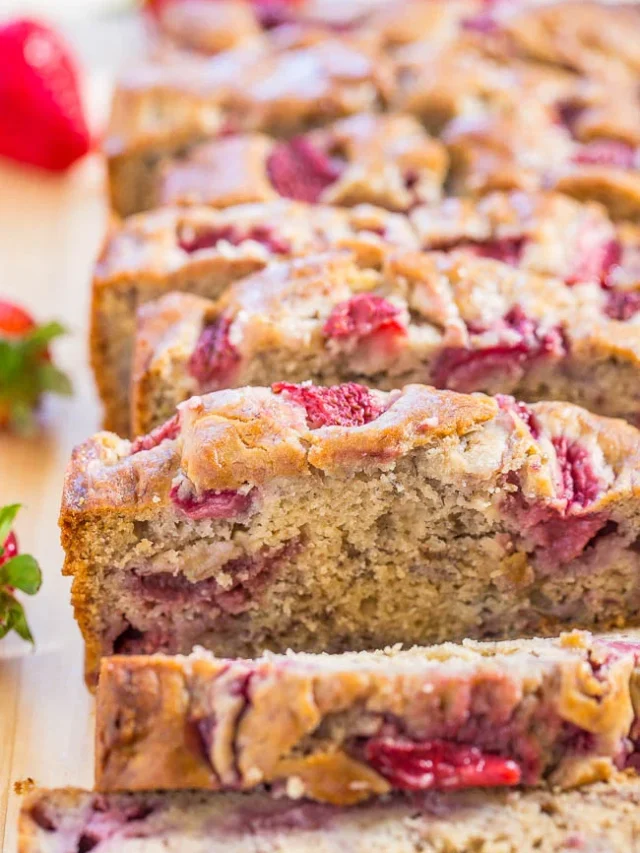 Sliced strawberry banana bread with visible chunks of strawberries on top, placed on a wooden surface. Some whole strawberries are visible in the background.
