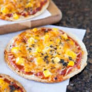 Three mini pizzas with cheese and diced yellow toppings on parchment paper and a wooden board.