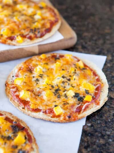 Three mini pizzas with cheese and diced yellow toppings on parchment paper and a wooden board.