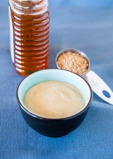 A bowl of tahini sauce with sesame seeds and a bottle of honey in the background.