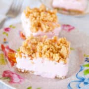 Two pink frosted dessert bars with crumb topping on a floral plate, with a fork in the background.