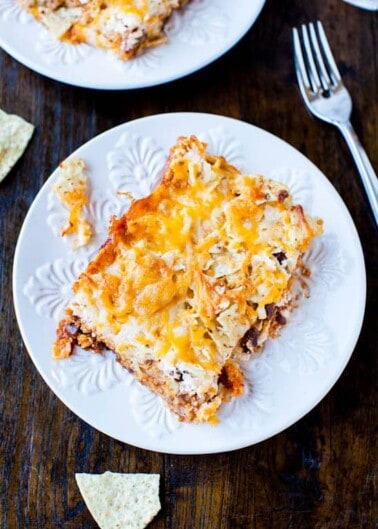 A slice of cheesy lasagna on a white plate with a fork to the side and a few scattered chips.