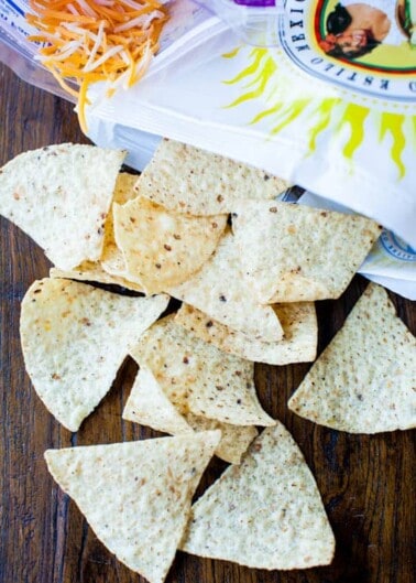 Tortilla chips scattered on a wooden surface next to a bag of shredded cheese and a container of sour cream.