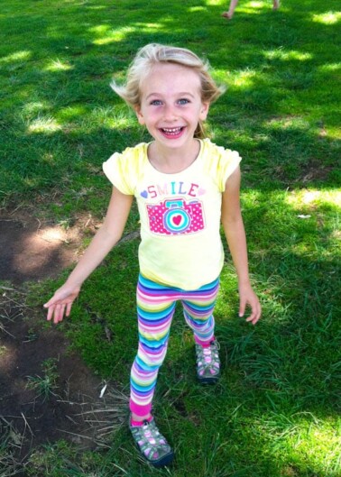 A smiling young girl in colorful striped leggings and a yellow t-shirt stands on grass.
