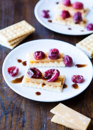 Cheese and crackers topped with raspberries and drizzled with a caramel-like sauce on white plates.