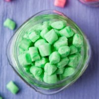 A glass jar filled with green mint-flavored chewing gum pieces on a purple surface.