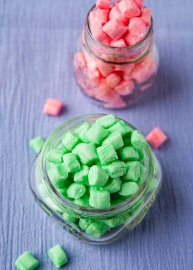Two jars of colored chewing gum, one with green pieces on a textile surface and another with pink pieces slightly out of focus in the background.