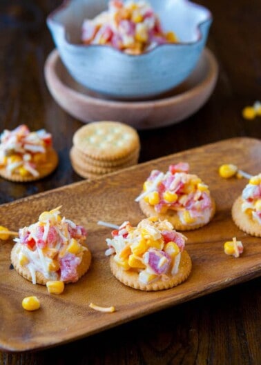 Crackers topped with a mixture of crab, corn, and cheese on a wooden board.