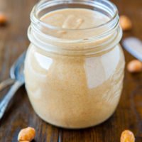 A jar of creamy peanut butter with scattered peanuts around it.