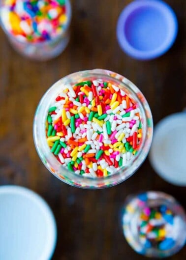 A jar of colorful sprinkles viewed from the top with additional jars and lids in the background.