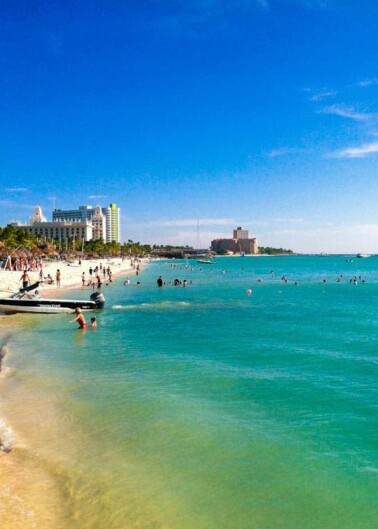 Beachgoers enjoying a sunny day on a tropical beach with clear blue waters and a skyline of hotels in the distance.
