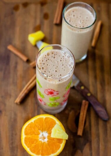 Two glasses of a creamy, frothy beverage garnished with cinnamon sticks and a sliced orange on a wooden surface.