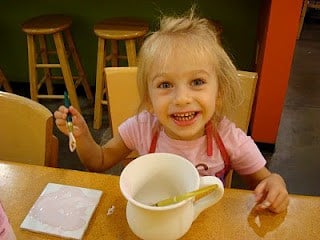 A young child smiling at a table with a paintbrush in hand and a bowl in front of her.