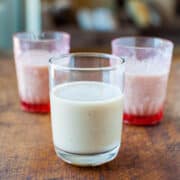 Three glasses on a wooden surface, one filled with a white creamy liquid and two with a pink liquid, possibly a beverage or dessert.