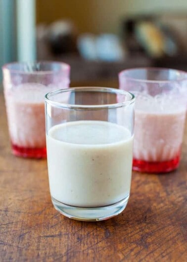 Three glasses on a wooden surface, one filled with a white creamy liquid and two with a pink liquid, possibly a beverage or dessert.