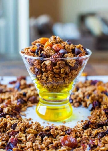 A glass filled with granola and mixed dried fruits, with scattered granola around the base on a table.