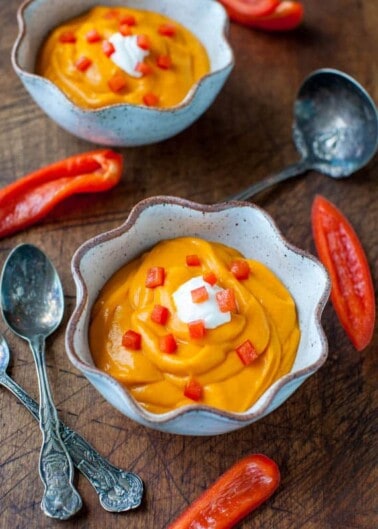Two bowls of creamy orange soup garnished with red pepper dices and a dollop of sour cream, accompanied by spoons and fresh pepper slices on a wooden surface.