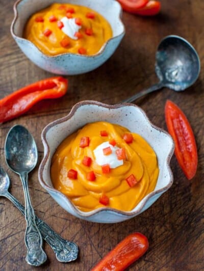 Two bowls of creamy orange soup garnished with red pepper dices and a dollop of sour cream, accompanied by spoons and fresh pepper slices on a wooden surface.